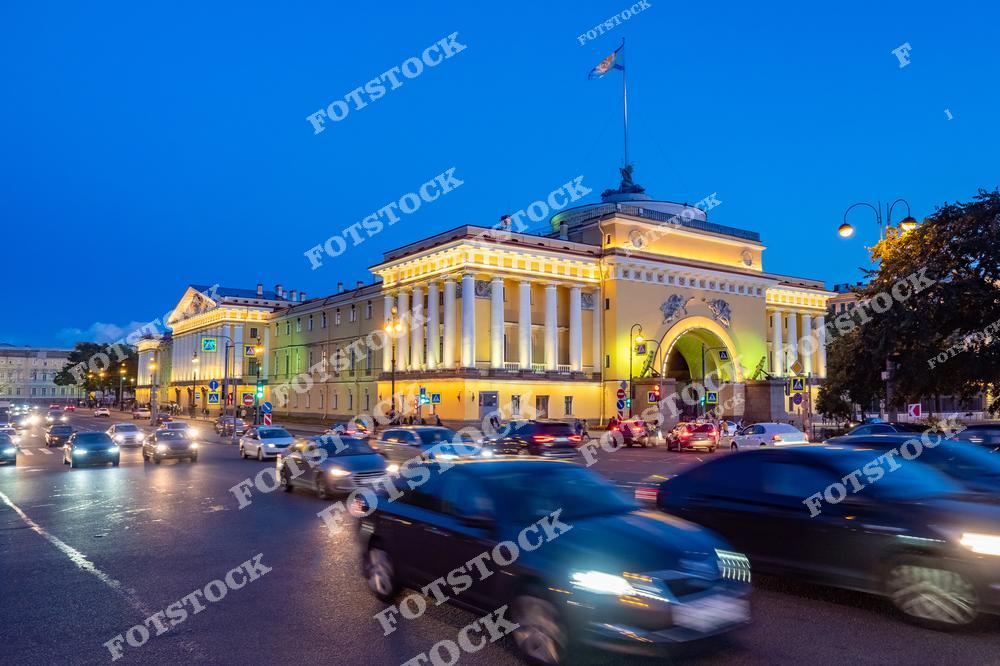На фотографии изображено Адмиралтейство — одна из главных достопримечательностей Санкт-Петербурга. Здание освещено вечерними огнями и выделяется на фоне насыщенного синего неба. Перед ним — оживлённое движение автомобилей, подчёркивающее современный ритм города. Архитектура здания с колоннадой и скульптурной композицией на крыше отражает величие имперского стиля. Это знаковое место, откуда традиционно начинается отсчёт всех главных улиц Петербурга.